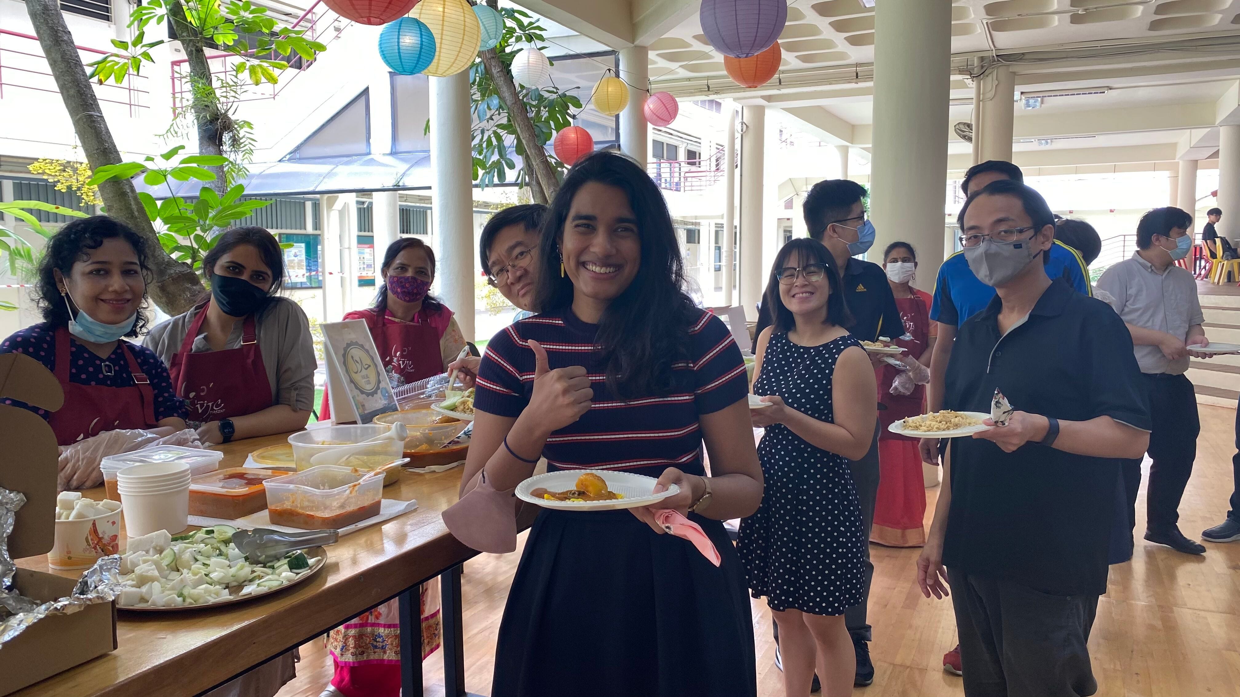 A smiling woman gives a thumbs up holding a food plate at an outdoor gathering with colorful paper lanterns overhead.